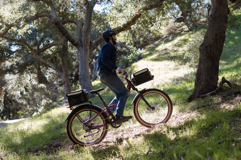 Image of a man riding the Radster Trail in Copper Red in a wooded area. He is wearing a helmet & has both a front-mounted basket & rear-mounted large basket on his bike. 