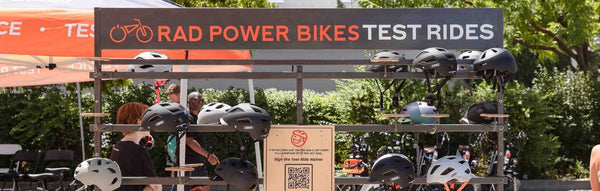 The upper half of a tent with the words Rad Power Bikes Test Rides on a sunny parking lot. Helmets are seen lined up below.