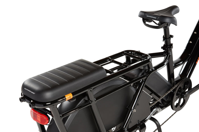 RadWagon deckpad installed on the back of a RadWagon electric cargo bike, used for passenger seating. Black vinyl material with padding. Subtle Rad Power Bikes logo along the end.