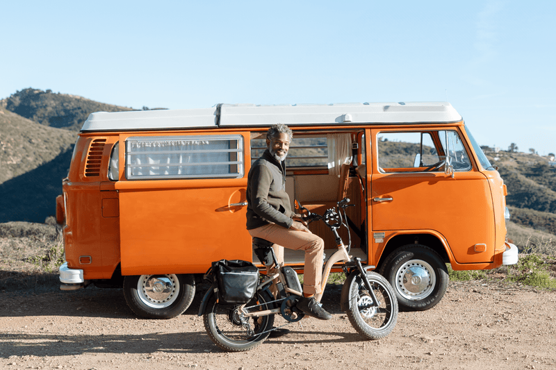 Image of man on RadExapnd 5 Plus in Stone Tan outside with a vintage camper van in the background. The scenary is outdoorsy.
