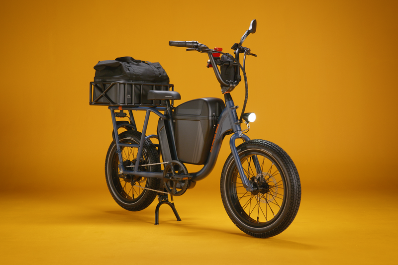 A blue cargo utility electric bike with a black kickstand, rear cargo basket, illuminated headlight, mirror, and storage console against a gold background.