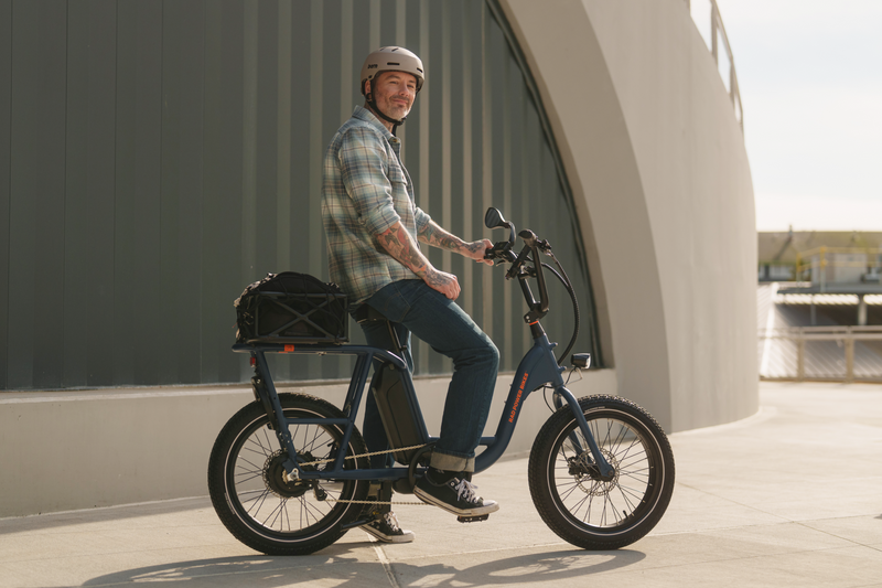 A man with tattoos wearing a plaid shirt, jeans, and a grey helmet smiling and sitting on an ebike in an urban setting. 
