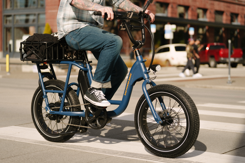 A man riding a blue electric bicycle with an orange Rad Power Bikes logo, fat black tires, and large basket with a cargo net, on a paved road in an urban setting. 