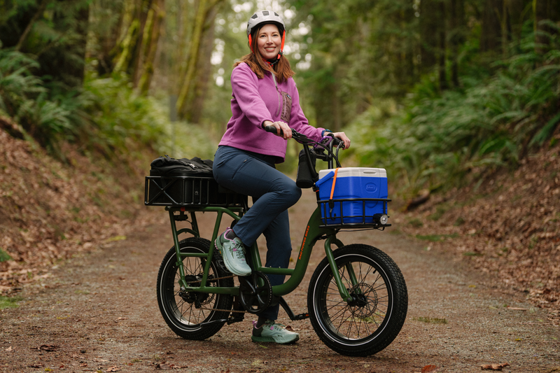 A woman on a trail in a forest sitting on a green cargo ebike with a small front basket and large rear basket wearing a pink jacket and white helmet.