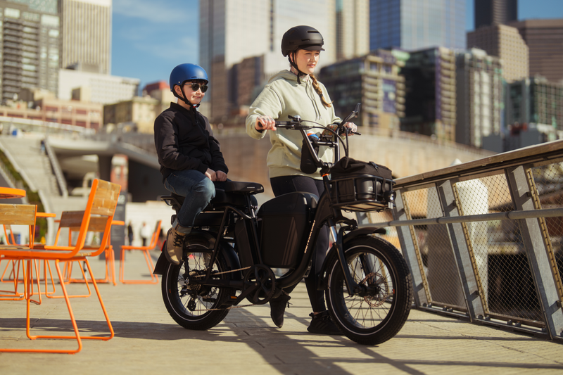 A woman wearing a tan hoodie and black helmet holding the handlebars of a matte black electric bike with a boy wearing a black shirt and jeans and blue helmet sitting on the back. On a bridge in an urban setting. 