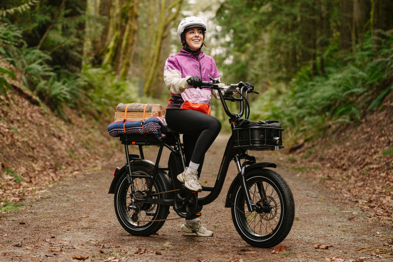 A woman sitting on a black RadRunner Plus electric cargo utility bike wearing a white helmet, pink jacket and grey pants. 