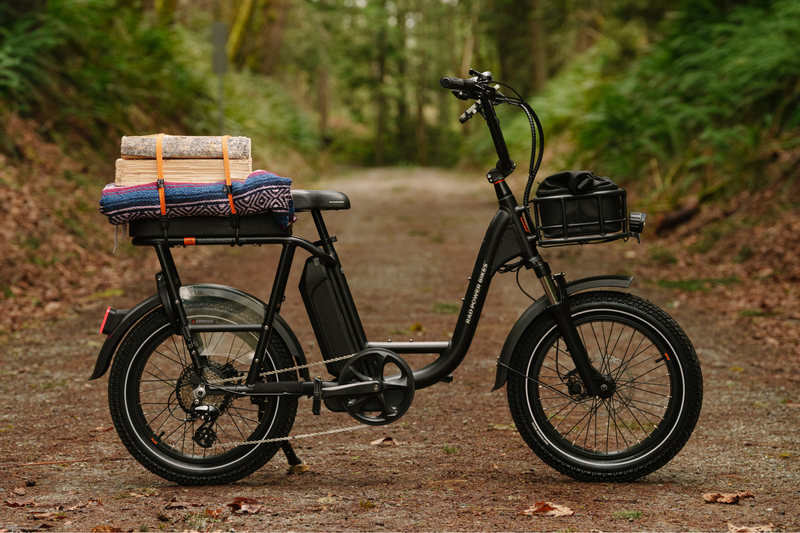 A black ebike on a dirt trail in a forest with a small basket on the front and wood and a blanket tied to the rear passenger seat. 